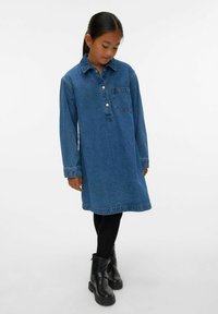 Jauna mergaitė stovi, vilkinti ilgomis rankovėmis džinsinę suknelę su priekiniais mygtukais, juodas pėdkelnes ir juodus aulinukus, prieš paprastą foną.