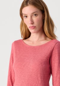 Garšām piedurknēm, apaļo izgriezumu ar mīkstu, teksturētu, izbalējušu rozā audumu. Iezīmē brīvu piegriezumu un vieglu materiālu ērtībai.