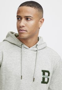 Light gray hoodie made of soft fabric, featuring a kangaroo pocket, drawstring hood, and green embroidered "B" logo with the word "BLEND."