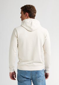 Cremefarbener Hoodie mit Kordelzugkapuze, langen Ärmeln und gerippten Bündchen, getragen mit blauen Jeans. Glatte Textur, minimale Details.