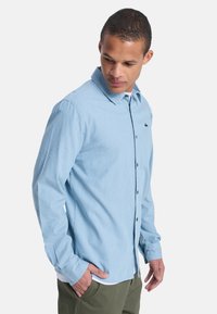 Hombre mirando hacia abajo, vistiendo una camisa de manga larga azul claro con botones y un pequeño logo en el pecho, y pantalones verde oliva.