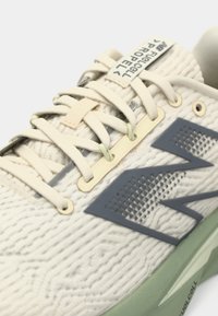 Hellbeige Sportchuhe mit strukturiertem Obermaterial, grauen Akzenten und einer grünen Sohle. Zu den Merkmalen gehören gewebte Schnürsenkel und ein Markenlabel.