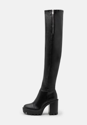 Botte en cuir noire montante jusqu’au genou avec talon bloc épais, semelle à crampons et fermeture éclair latérale sur fond uni.