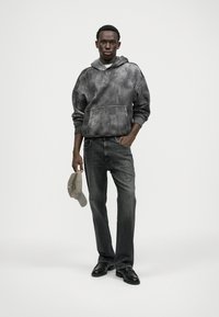 Γκρίζα φούτερ με tie-dye και μπροστινή τσέπη, συνδυασμένο με σκούρα γκρίζα τζιν με ίσια πόδια. Κρατάει ένα μπεζ καπέλο μπέιζμπολ. Μαύρα παπούτσια ολοκληρώνουν το ντύσιμο.