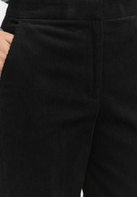 Zwarte corduroy broek met verticale ribbels, voorzien van zijzakken en een soepel tailleband. De stof heeft een zachte, gestructureerde uitstraling.