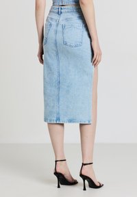 Femme portant une jupe midi en denim bleu clair avec des poches arrière et une fente sur le côté, associée à des sandales à talons noirs, se tenant sur un sol neutre.