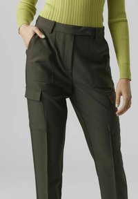 Olivengrüne Hose mit Seitentaschen, taillierte Passform und glatter Textur. Kombiniert mit einem ribbeligen, limegrünen Pullover.