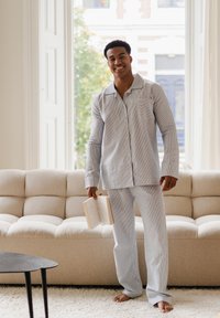 Ensemble de pyjama rayé bleu clair et blanc, composé d'une chemise à manches longues avec boutons et poche poitrine, assortie d'un pantalon, sur un canapé beige.