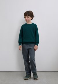 Suéter tejido color azul verdoso con puños acanalados, combinado con pantalones grises texturizados y zapatillas verdes con cierres de tira. Fondo liso.