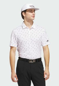 White polo shirt with a pattern of small black triangles, paired with black trousers and a white cap featuring the text "ADIDAS" in black.