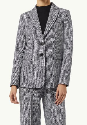 Kvinde iført en sort og hvid blazer med fiskebensmønster og matchende bukser over en sort rullekravesweater.