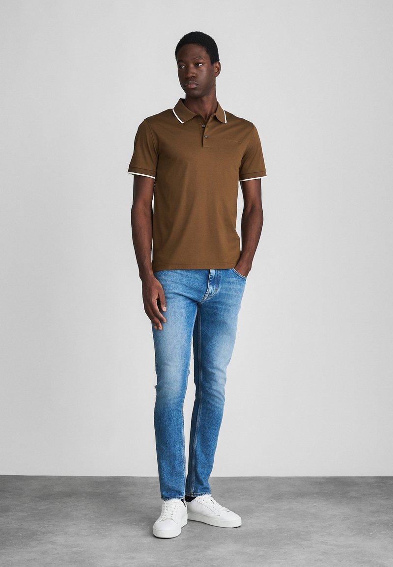 Camisa polo marrón con cuello y detalles en blanco, combinada con jeans de color azul claro y zapatillas blancas. El modelo está de pie contra un fondo neutral.