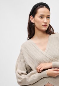 Pulover bej înfășurat, cu textură canelată, decolteu adânc în V și mâneci largi. Modelul poartă o bandă de cap din mărgele negre și cercei cu cercuri din argint.