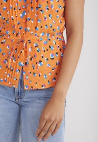 Kortärmad, orange topp med ett samlat mönster. Har flerfärgade prickar, knappar och en knytning i midjan. Bärs med blå jeans.