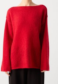 Persona che indossa un maglione rosso peloso oversize a maniche lunghe con scollo ampio e pantaloni neri su uno sfondo bianco.