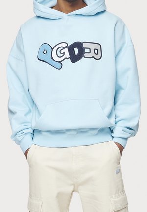 Personne portant un sweat à capuche bleu clair avec des lettres texturées et ludiques "PGDEO" sur le devant, et un pantalon cargo beige les mains dans les poches.