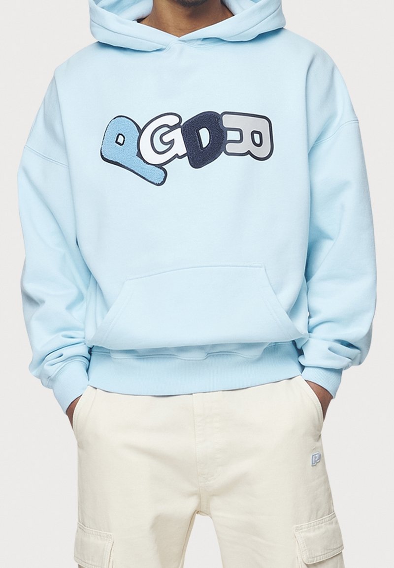 Personne portant un sweat à capuche bleu clair avec des lettres texturées et ludiques "PGDEO" sur le devant, et un pantalon cargo beige les mains dans les poches.