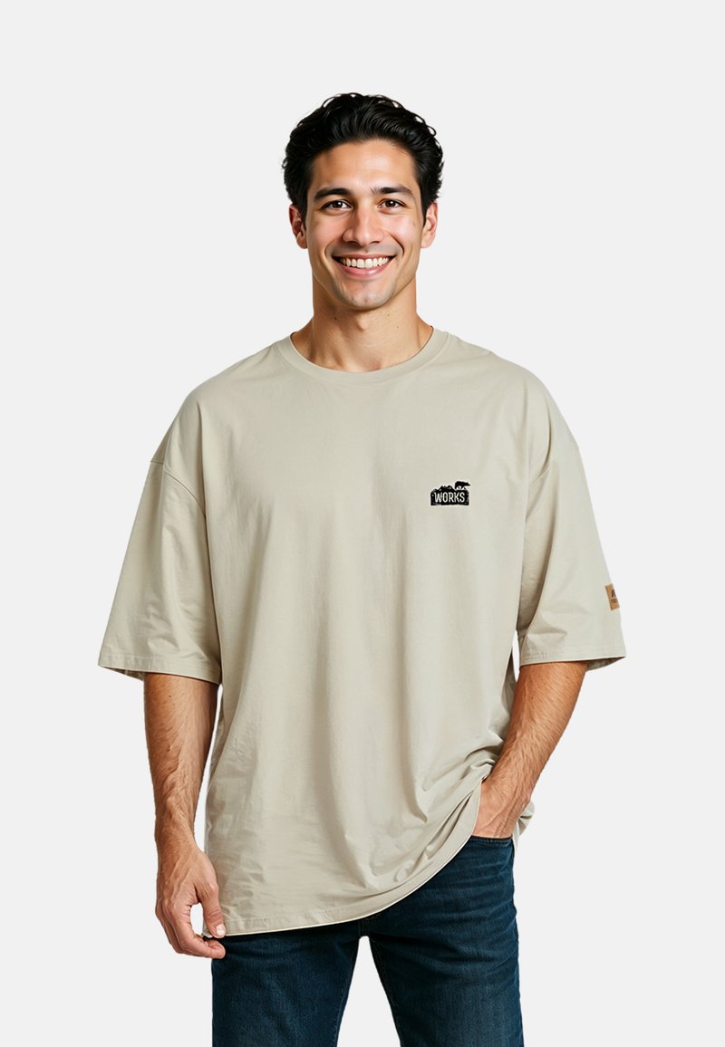 Beige Übergröße T-Shirt mit kurzen Ärmeln, rundem Ausschnitt und einem kleinen schwarzen Logo auf der Brust. Kombiniert mit dunkelblauen Jeans.