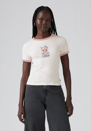 Kvinna med flätat hår som bär en krämfärgad t-shirt med rosa detaljer och en tecknad kattgrafik, i kombination med högmidjade mörkgrå vida byxor.