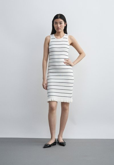 Γυναίκα στέκεται με το ένα χέρι στη μέση, φορά ένα λευκό αμάνικο φόρεμα με λεπτές μαύρες οριζόντιες ρίγες και μαύρα μυτερά παπούτσια χωρίς τακούνι.