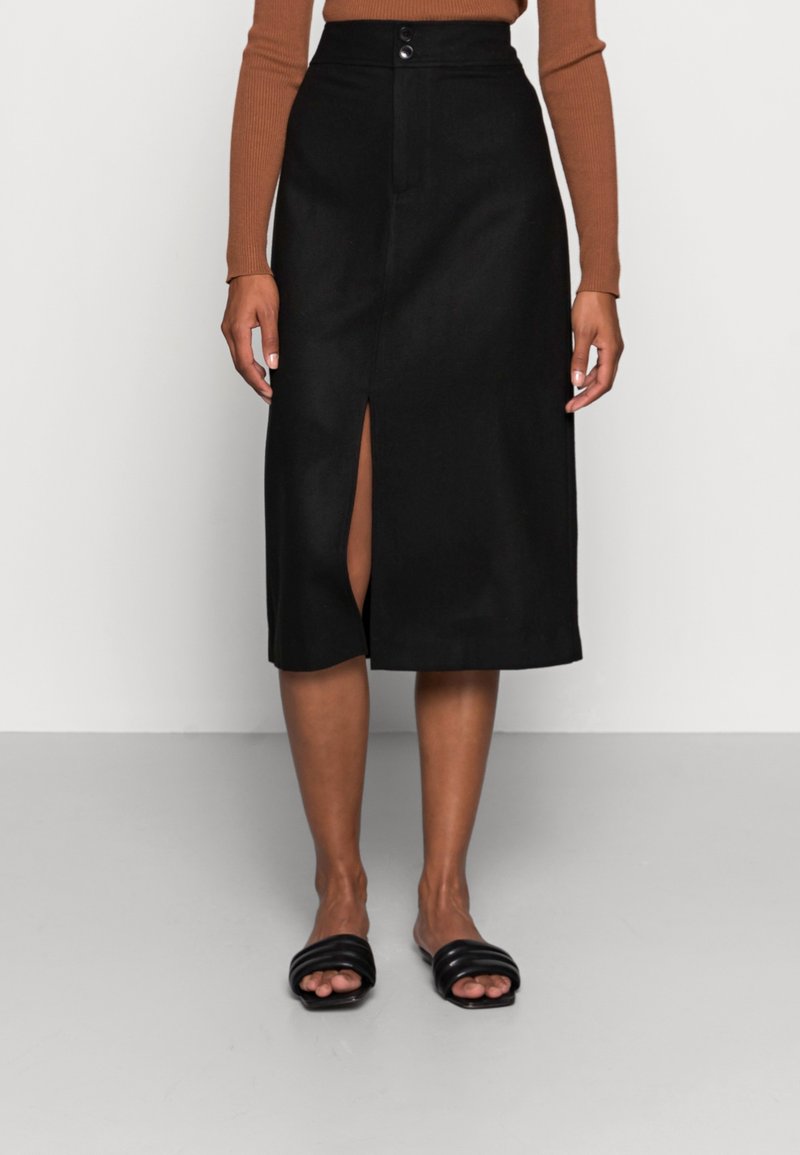 Femme portant une jupe crayon noire longueur genoux avec une fente frontale, un haut à manches longues marron, et des sandales slides noires rembourrées, debout sur un sol gris.