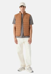 Gilet imbottito marrone con cuciture verticali, abbinato a una t-shirt grigia e pantaloni bianchi. Il modello indossa un berretto chiaro e scarpe da ginnastica grigie.