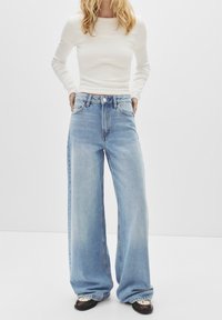 Jeans bleu clair, à jambes larges, taille haute, avec cinq poches et une fermeture à bouton. Associés à un haut ajusté à manches longues blanc et des chaussures noires.
