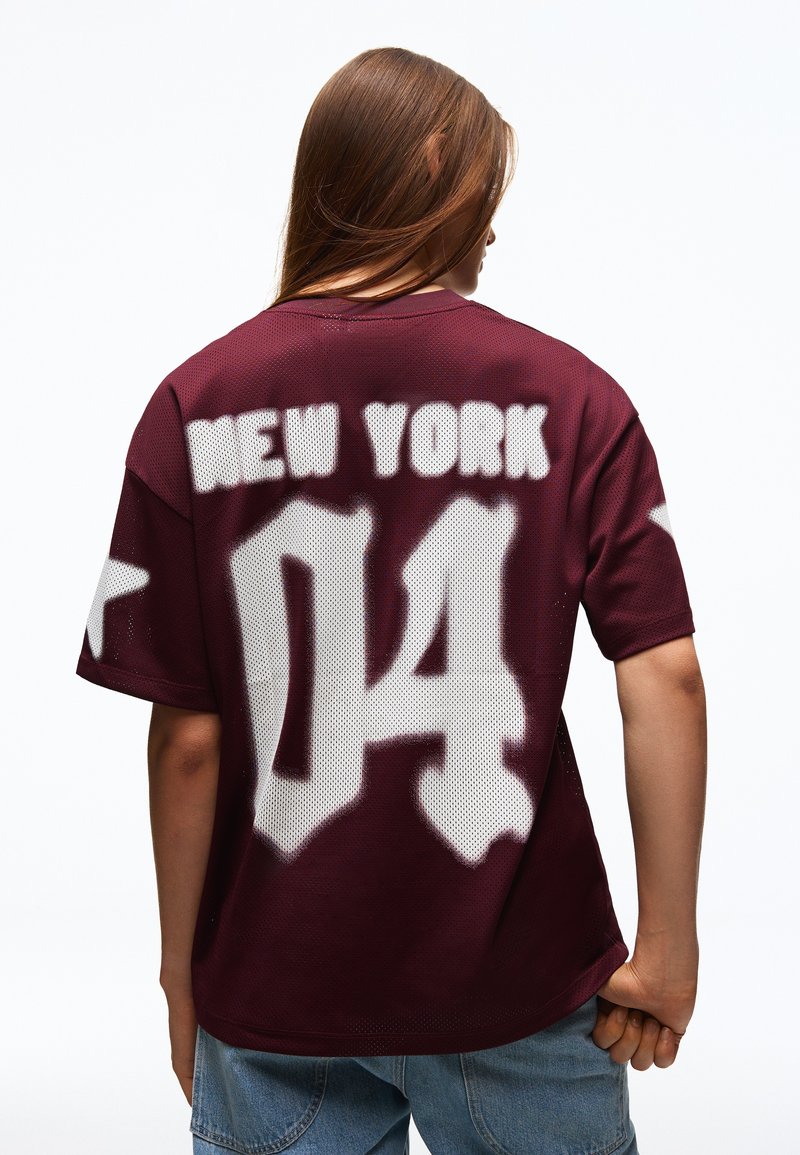 Personne aux longs cheveux bruns portant un maillot bordeaux avec des étoiles blanches sur les manches et un grand texte "NEW YORK 04" dans le dos.