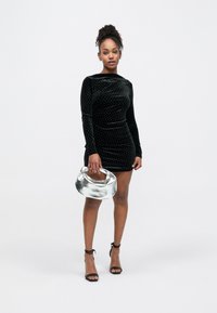 Rochie din catifea neagră cu buline argintii, mâneci lungi și croială conică. Accentuată cu o geantă metalică argintie și sandale negre cu barete.