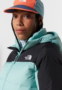 Podstavljena jakna u tirkiznoj i crnoj boji, s patentnim zatvaračem sprijeda, kapuljačom i logotipom The North Face. Šarena kapa upotpunjuje ovaj outfit.