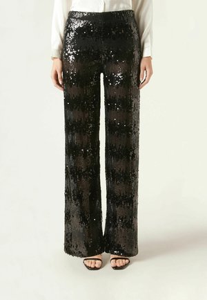 Donna che indossa pantaloni neri a vita alta e gamba larga coperti da paillettes nere lucide, abbinati a una camicia bianca e sandali neri con punta aperta.