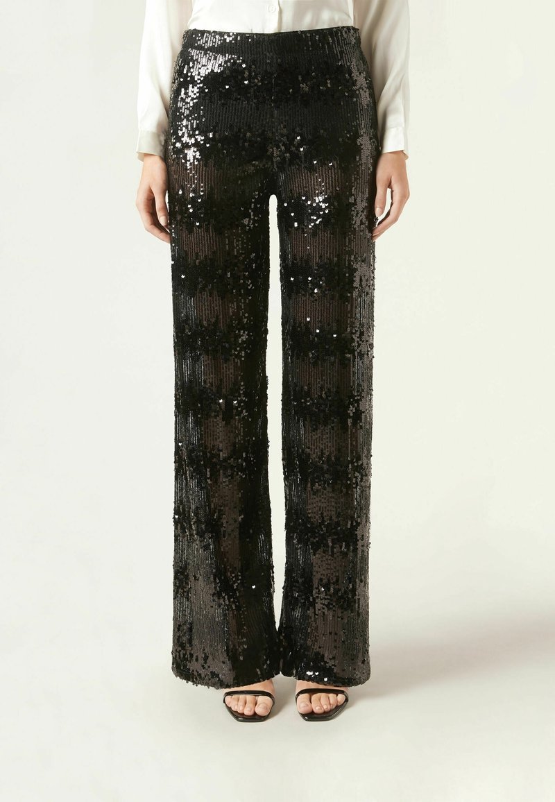 Femme portant un pantalon noir taille haute à jambes larges recouvert de sequins noirs brillants, associé à une chemise blanche et des sandales noires ouvertes.
