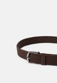 Vävd brun bälte med en silverfärgad metallspänne, med en strukturerad mönster och en läderdetalj med en logotyp i änden.
