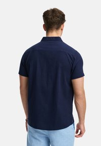 Camisa de manga corta en tejido texturizado azul marino, con cuello, corte recto y dobladillo curvado. La vista trasera muestra un diseño minimalista.