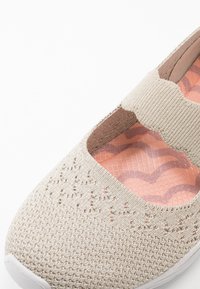 Zapato beige tipo slip-on hecho de material de punto con una correa ondulada. La parte superior texturizada presenta pequeños recortes. El interior tiene un forro patrón en color rosa.