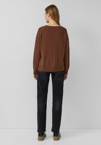 Brązowy dzianinowy sweter z prążkowanym ściągaczem u dołu i przy szyi, w połączeniu z czarnymi jeansami o prostych nogawkach i czarnymi botkami na masywnej podeszwie. Neutralne tło.