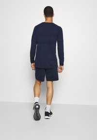 Hombre de pie frente a una pared, vistiendo una camisa de manga larga azul marino, pantalones cortos azul marino, calcetines blancos y zapatos deportivos negros con suelas blancas.