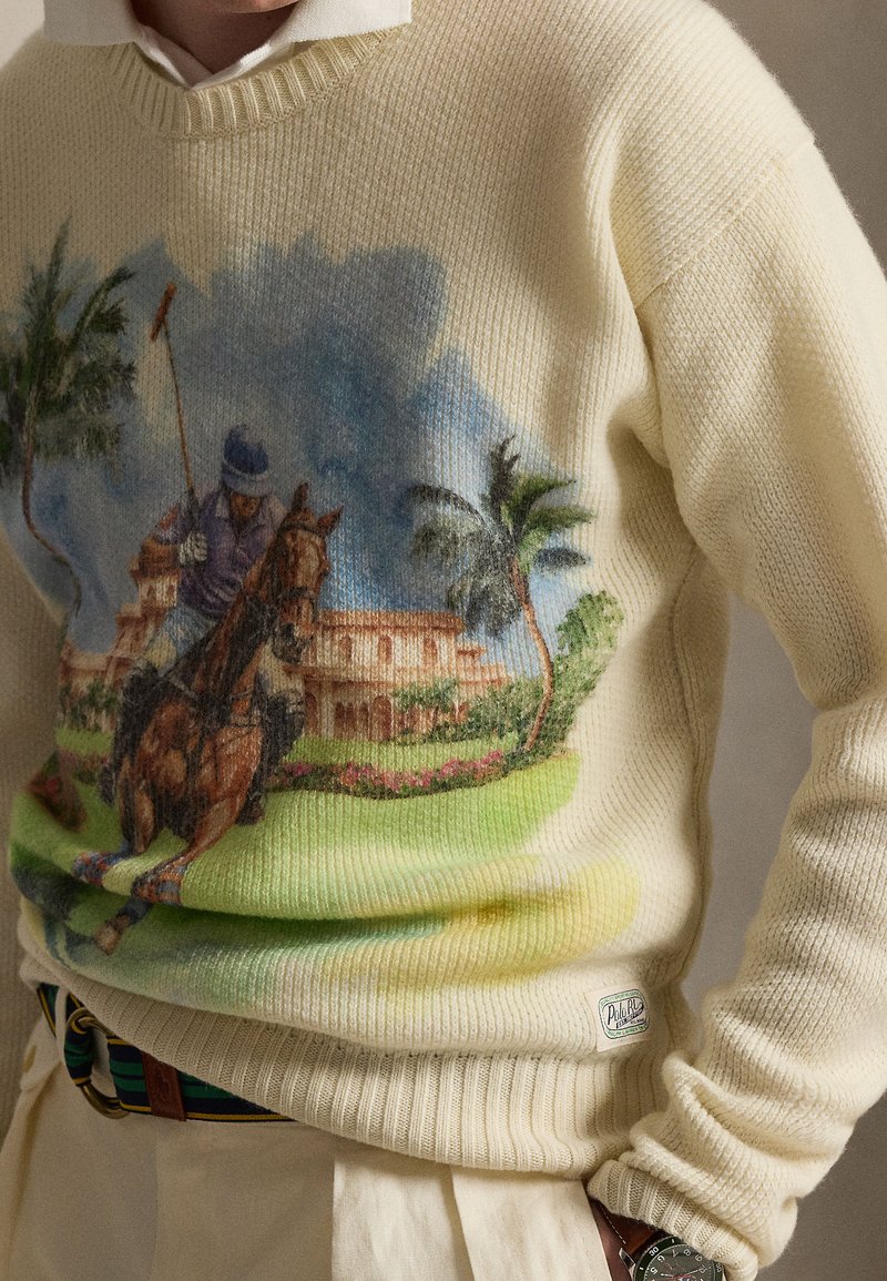 Maglione crema con una stampa dettagliata di un giocatore di polo a cavallo davanti a una villa e palme, indossato sopra una camicia bianca.
