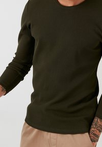 Top a maniche lunghe, verde oliva, in maglia a costine con scollatura rotonda e maniche aderenti, che mette in evidenza una superficie testurizzata e una leggera elasticità.