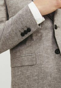 Blazer de lino gris claro con un acabado texturizado. Presenta tres botones negros en el puño y un bolsillo en el pecho.