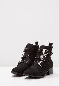 Botas de tobillo de ante negro con tres correas de hebilla plateadas decoradas con pequeños puntos plateados, tacones de bloque y cremalleras laterales.