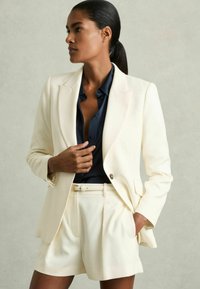 Completo giacca e pantaloni corti crema, con chiusura a un bottone, rever a lancia e tasche laterali. Indossato sopra una camicia blu scuro.