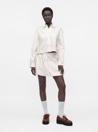 Femme portant une chemise à manches longues et une jupe assorties, claires et à motifs, des chaussettes blanches jusqu’aux mollets et des chaussures compensées marron, debout sur un fond uni.