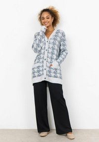 Cardigan houndstooth grigio e bianco con maniche lunghe, bottoni frontali, tasche e design a V, abbinato a pantaloni neri e scarpe chiare.