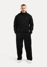 Muž stojící s rukama v kapsách, oblečený v černé mikině s krátkým zipem, odpovídajících černých kalhotách a bílých teniskách na bílém pozadí.