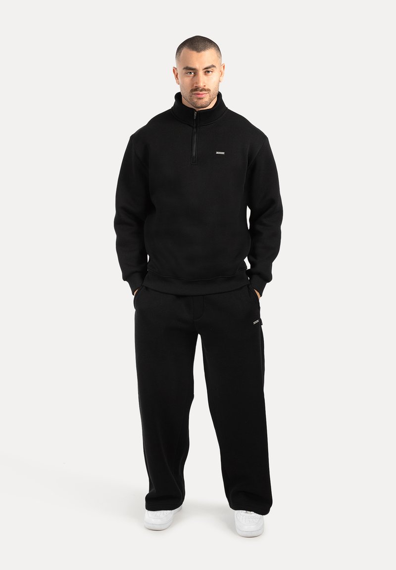 Muž stojící s rukama v kapsách, oblečený v černé mikině s krátkým zipem, odpovídajících černých kalhotách a bílých teniskách na bílém pozadí.