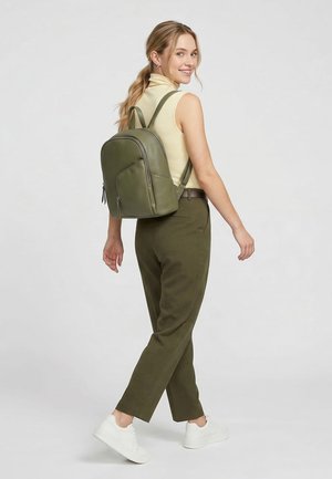 Donna sorridente che indossa una canotta beige e pantaloni color oliva, che cammina con uno zaino verde oliva e scarpe da ginnastica bianche su uno sfondo grigio uniforme.