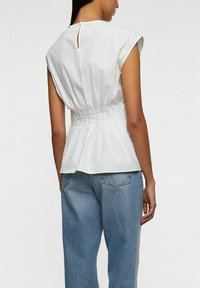 Hvid kortærmet bluse med en tætsiddende talje og plisseret ryg, som har en nøglehulsudskæring og en glat bomuldsstruktur. Samlet med lyseblå jeans.