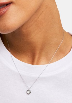 Silver chain necklace featuring a small, heart-shaped pendant with a clear gemstone, worn above a white shirt.