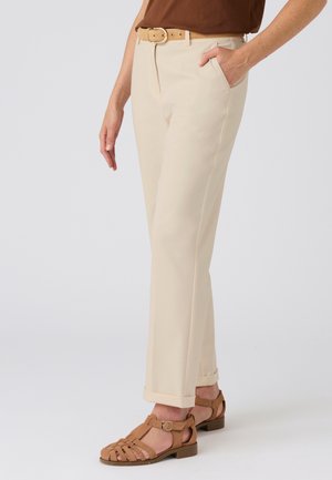 Pantaloni beige con vestibilità su misura, dotati di cintura, tasche e risvolti arrotolati. Indossati con sandali marroni con cinturini. Tessuto dalla consistenza liscia.